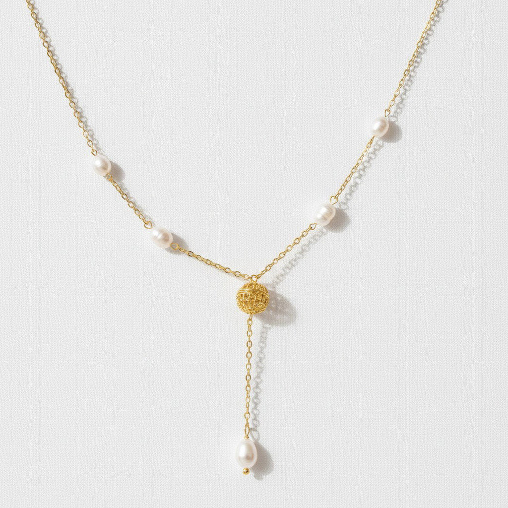 A delicate gold Y-necklace featuring small white pearls spaced along the chain and a textured gold circular pendant at the center, with a longer drop chain ending in a larger pearl, shown on a light gray background.