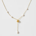 A delicate gold Y-necklace featuring small white pearls spaced along the chain and a textured gold circular pendant at the center, with a longer drop chain ending in a larger pearl, shown on a light gray background.