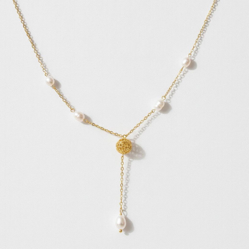 A delicate gold Y-necklace featuring small white pearls spaced along the chain and a textured gold circular pendant at the center, with a longer drop chain ending in a larger pearl, shown on a light gray background.
